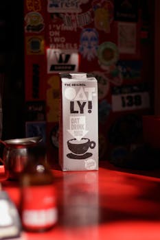 A carton of Oatly oat drink illuminated by sunlight on a red cafe counter, surrounded by eclectic decor.