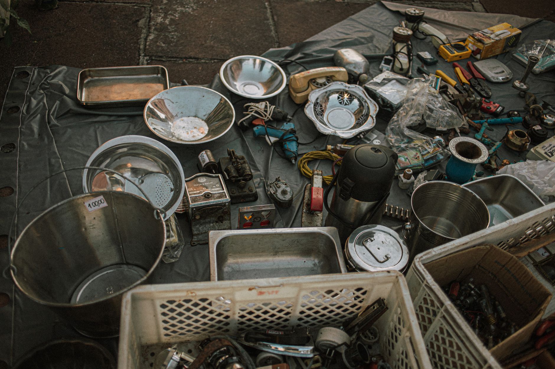 Assorted metal items and vintage tools displayed at an outdoor flea market on asphalt.