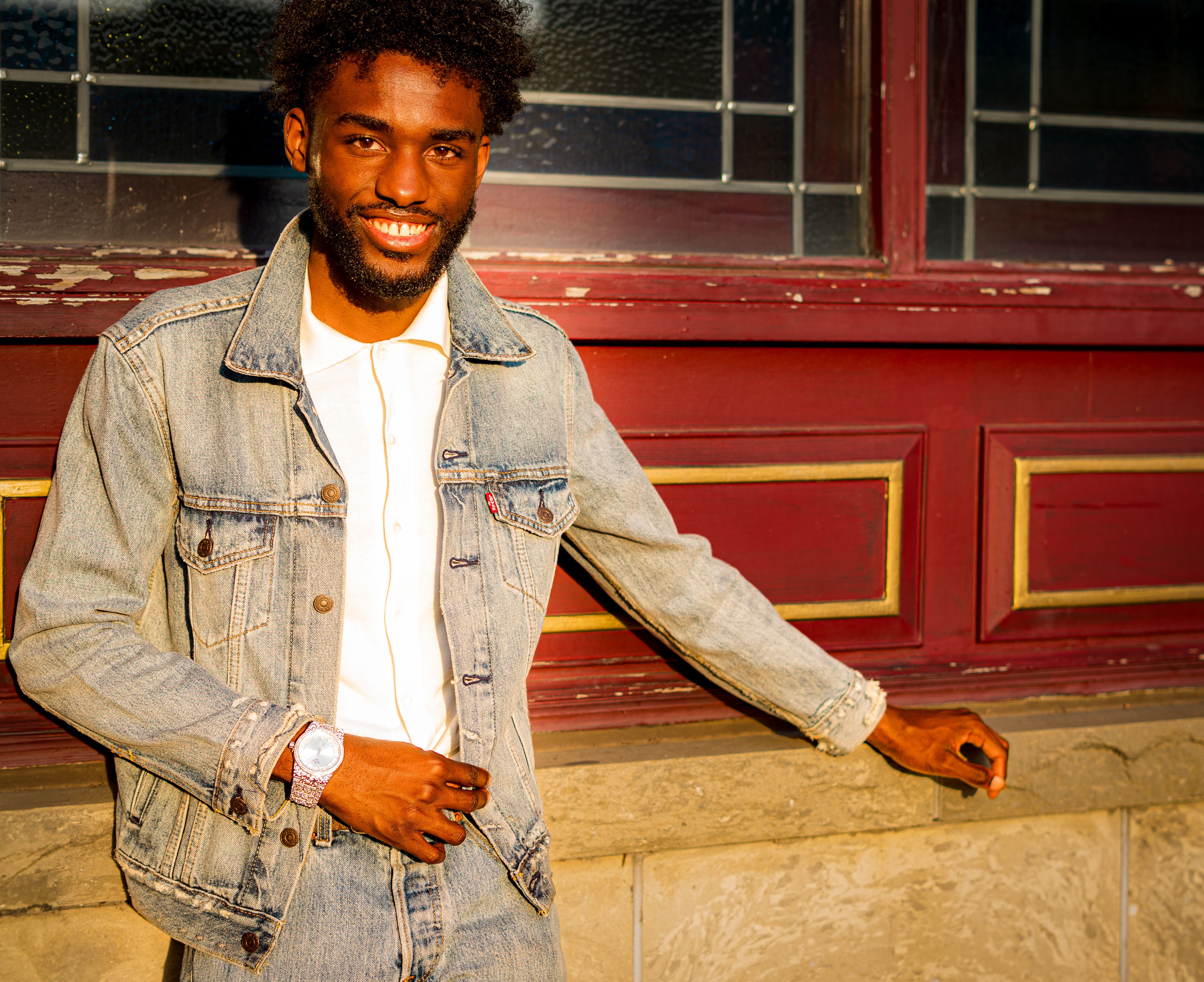 Smiling man wearing a denim jacket, standing against a decorated wall in warm sunlight.
