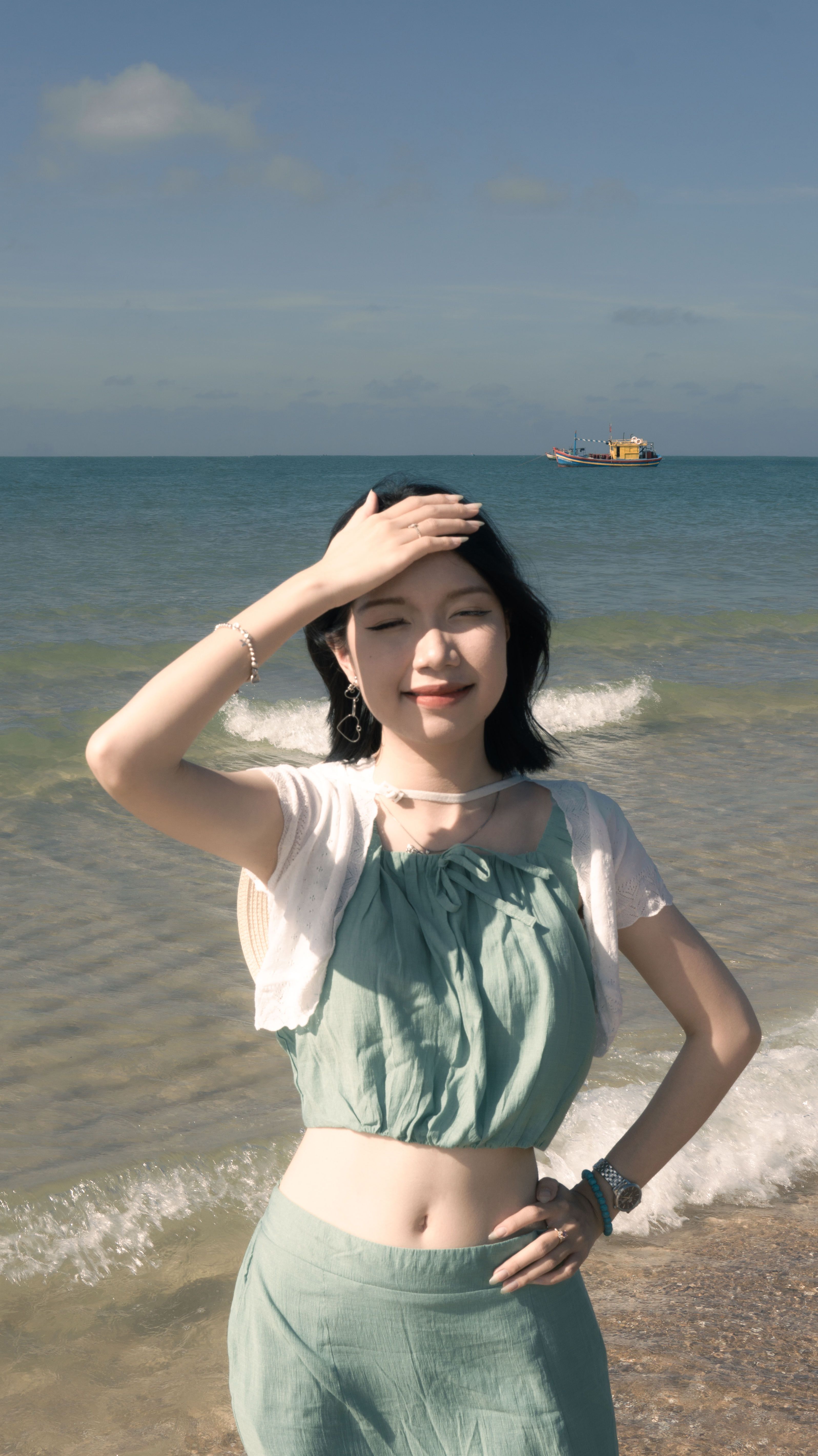 Smiling woman in green dress enjoys a sunny day at the beach, with waves and a distant boat.