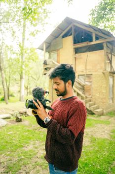 A young man filming outdoors in Badgam with a DSLR camera. Rustic backdrop, green foliage. - Fotografer Sidoarjo