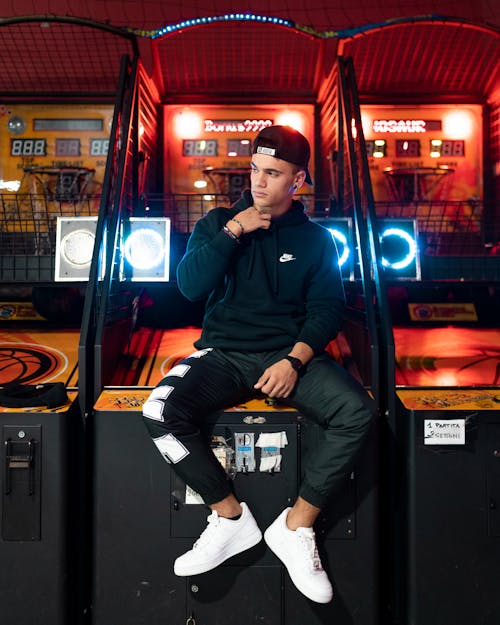 Full length calm young male wearing street style clothing sitting lordly on gaming machine and looking away with attitude