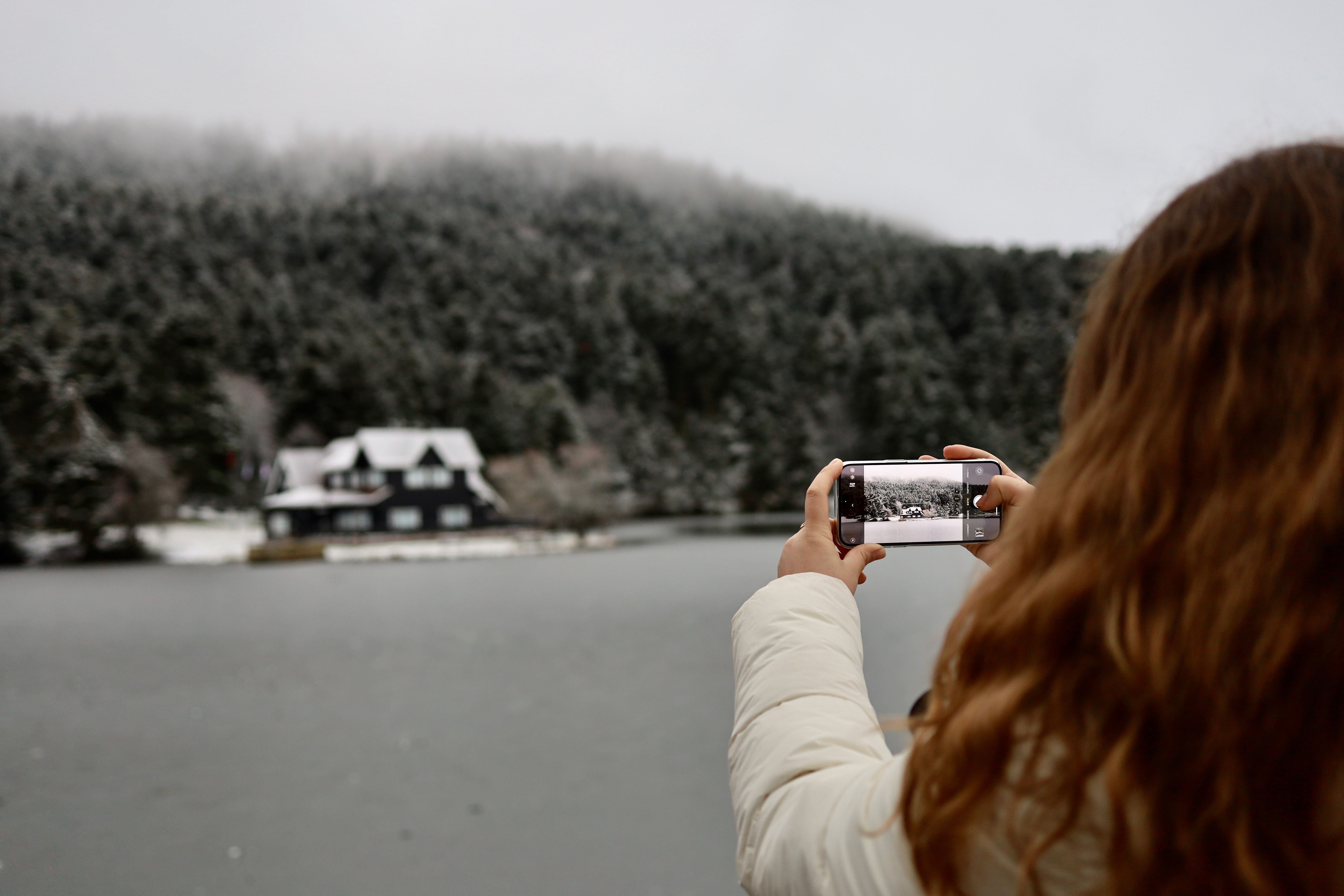 A woman photographs a snowy landscape and cottage at Lake Abant in Bolu, Turkey using a smartphone.