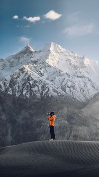 A lone photographer in an orange jacket captures a panoramic view of snowy mountain peaks.