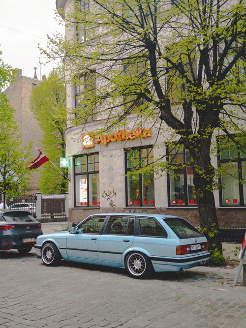 Free A classic blue station wagon parked outside a European pharmacy with lush spring foliage. Stock Photo