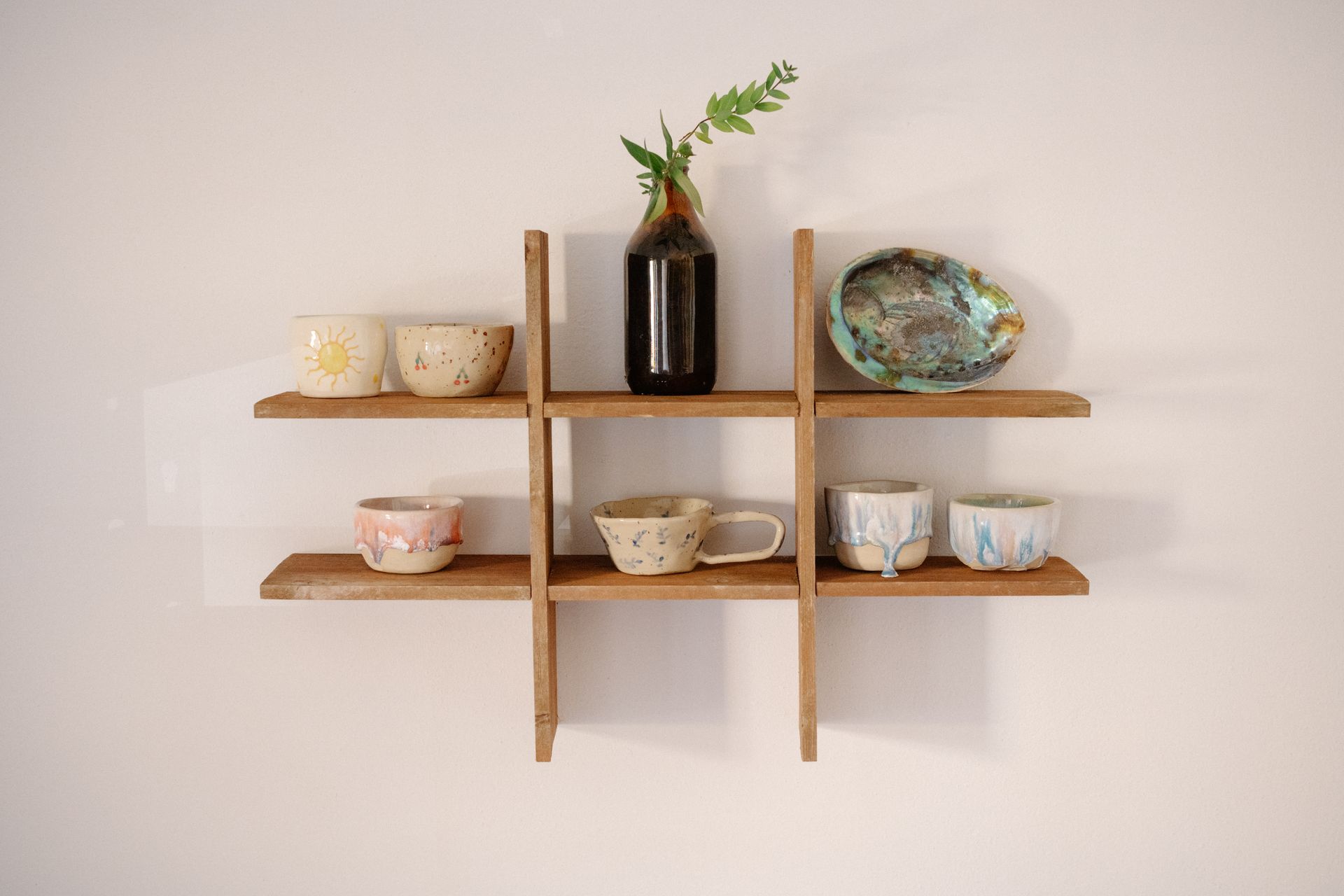 A wooden shelf displaying various handcrafted ceramic cups and vase in a minimalist style.