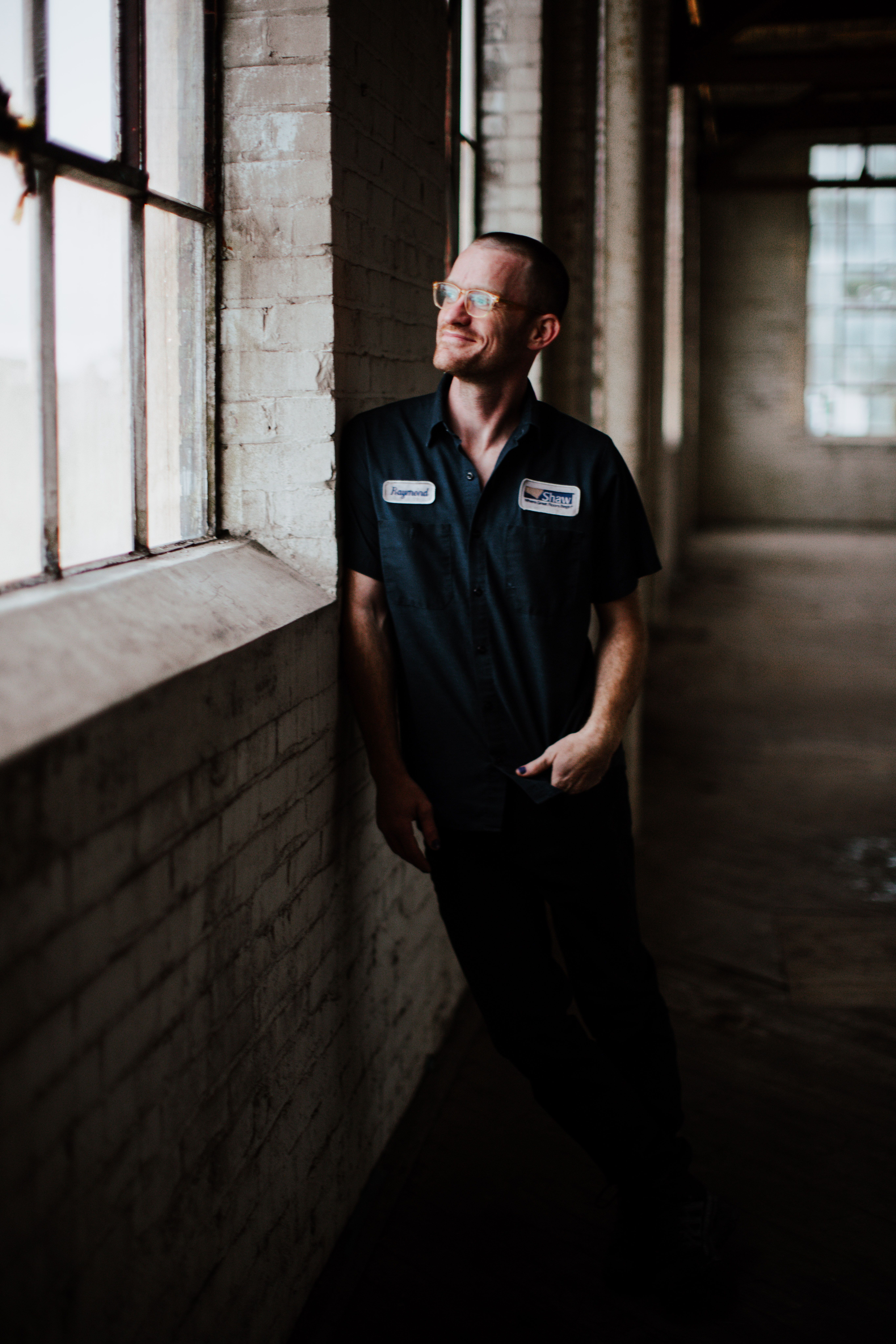 A man in a dark work shirt gazes thoughtfully outside through a window in an industrial setting.
