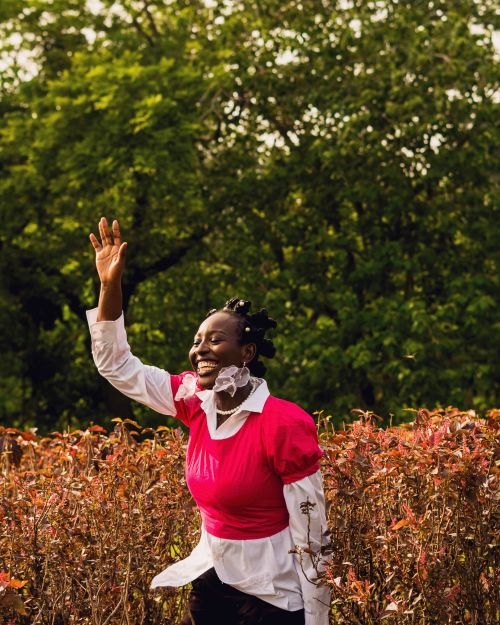 Free A woman dressed in bright clothing waves joyfully amidst a garden with autumn foliage. Stock Photo