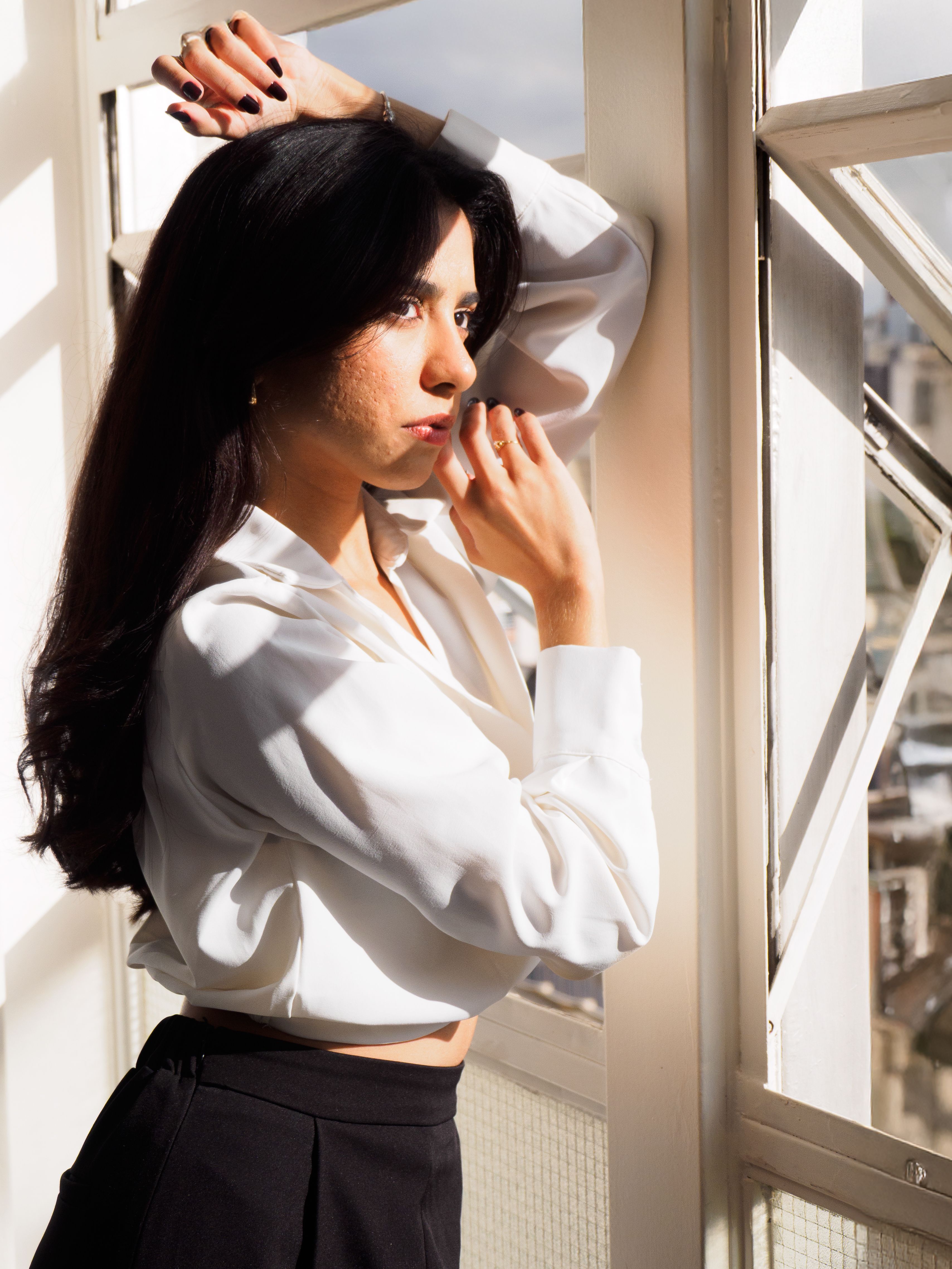 A thoughtful woman in a white shirt gazes out a bright window, bathed in sunlight.