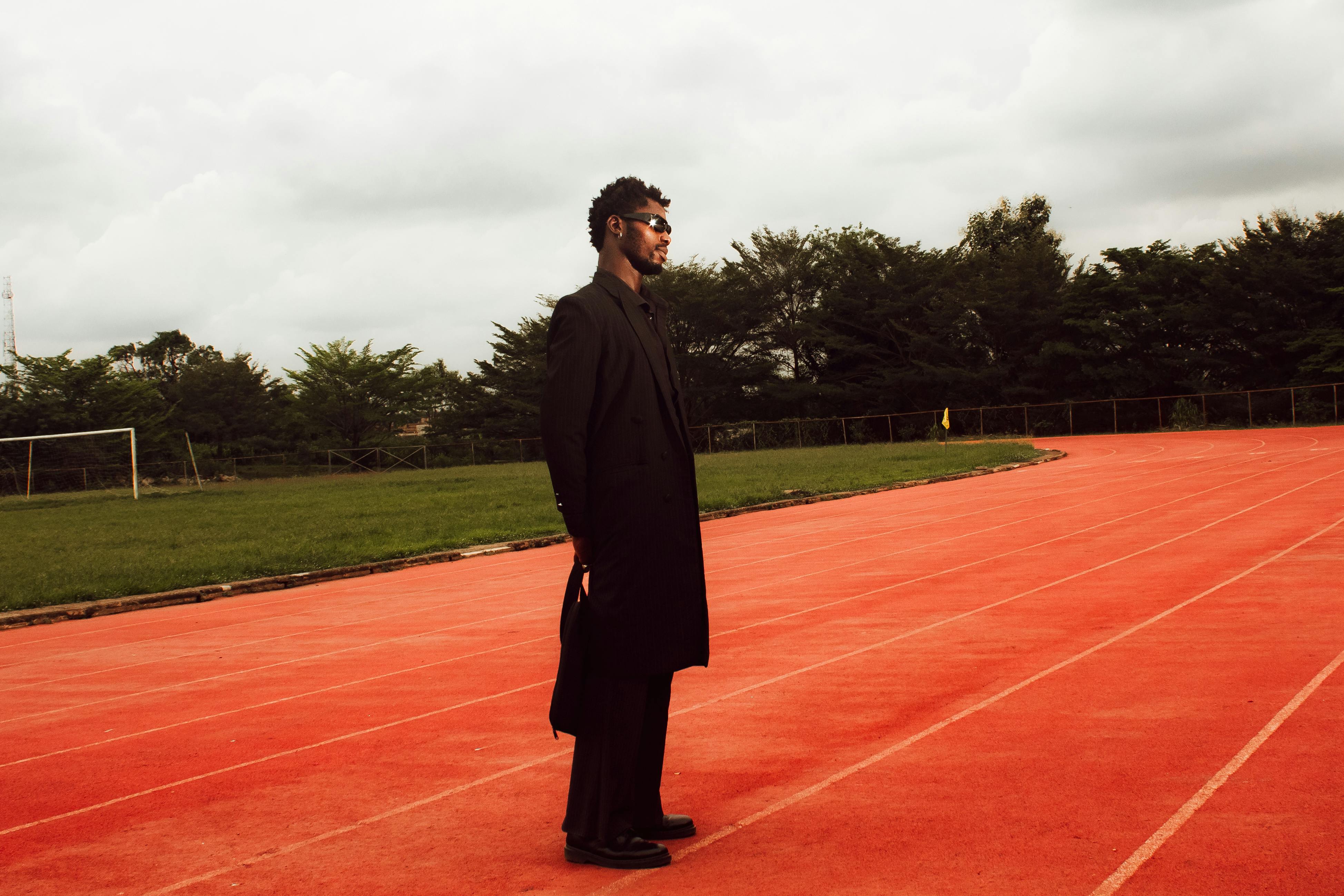 A stylish man in black clothing stands confidently on a red running track outdoors.