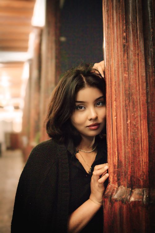 Free Elegant woman leaning against a wooden pillar indoors, soft lighting. Stock Photo