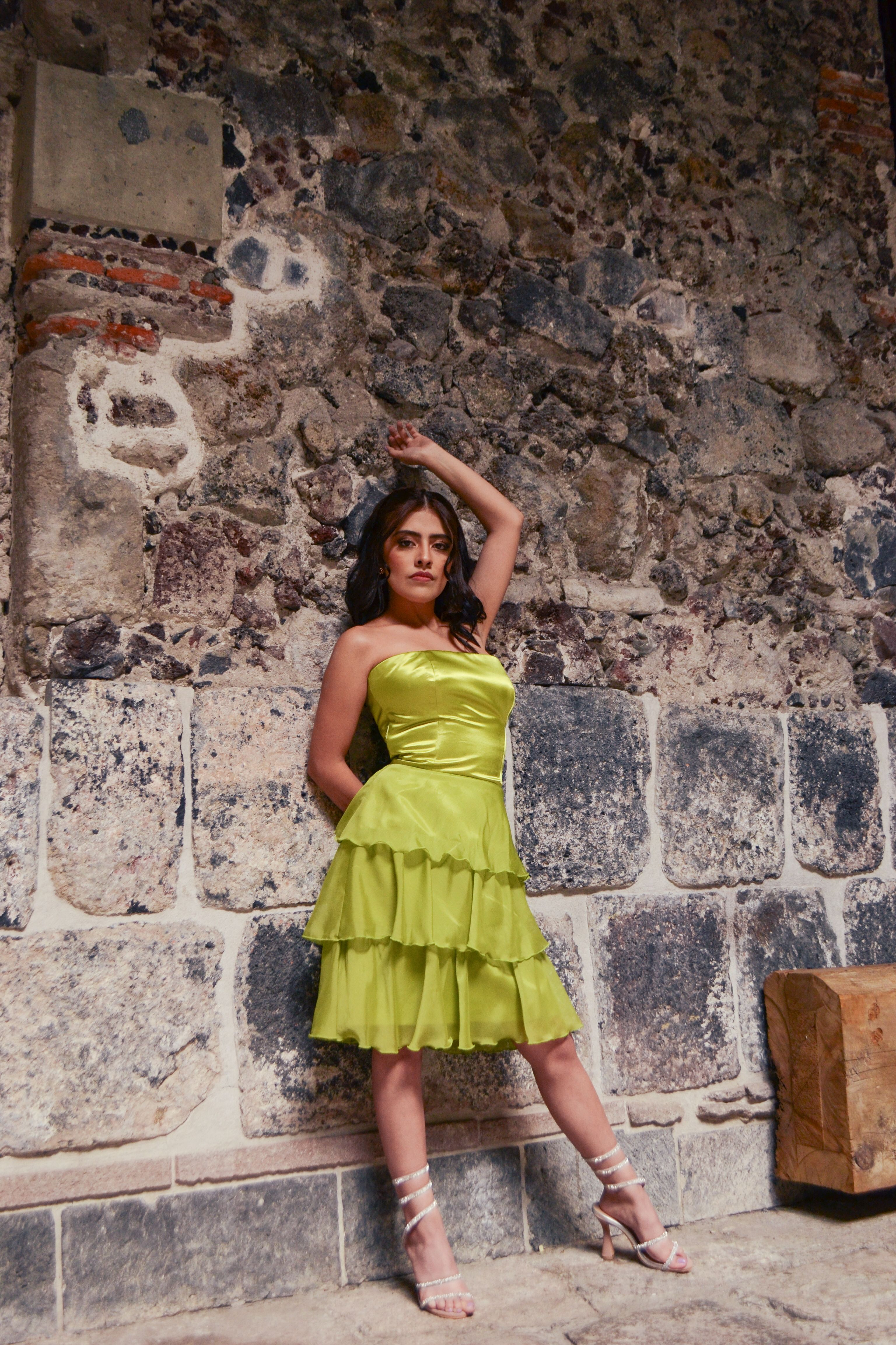 A stylish woman in a lime green dress poses against a rustic stone wall in an outdoor setting.