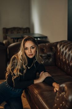 Stylish woman with long hair and necklace posing on brown leather sofa in dimly lit room.