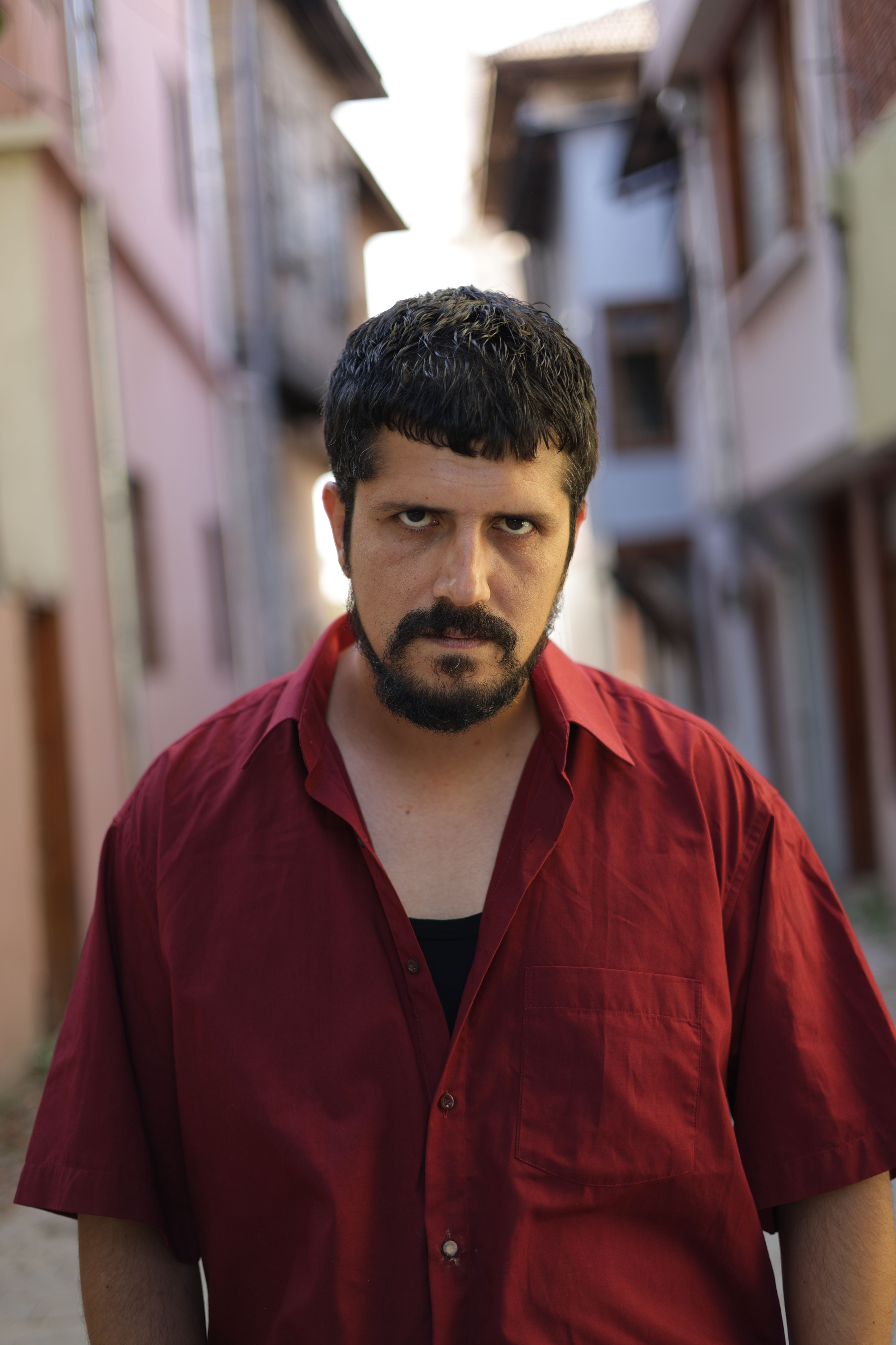 A serious-looking man in a vibrant red shirt stands in a narrow alleyway in Türkiye.