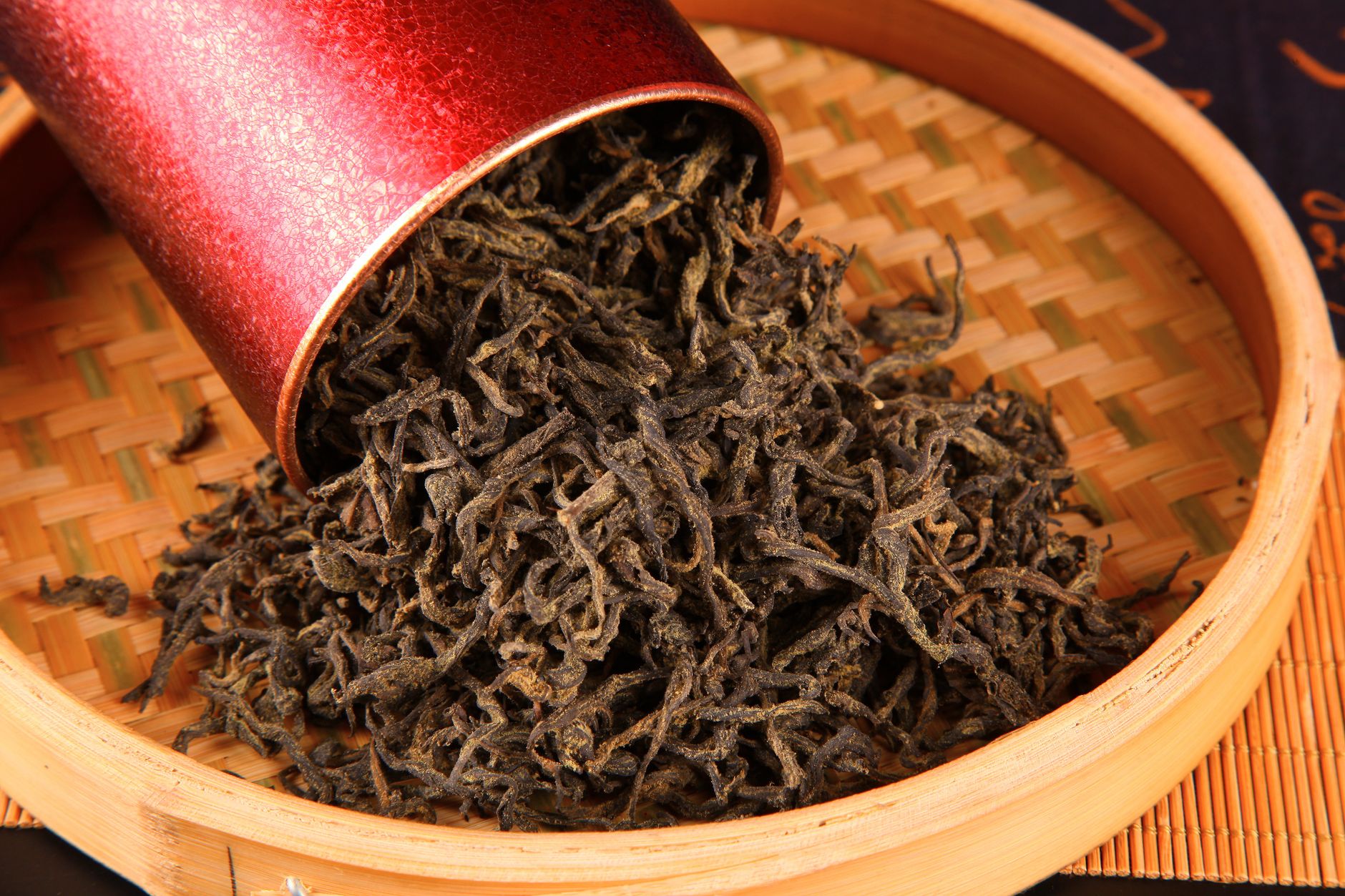 Close-up of loose black tea leaves spilling from a red canister onto a woven tray.