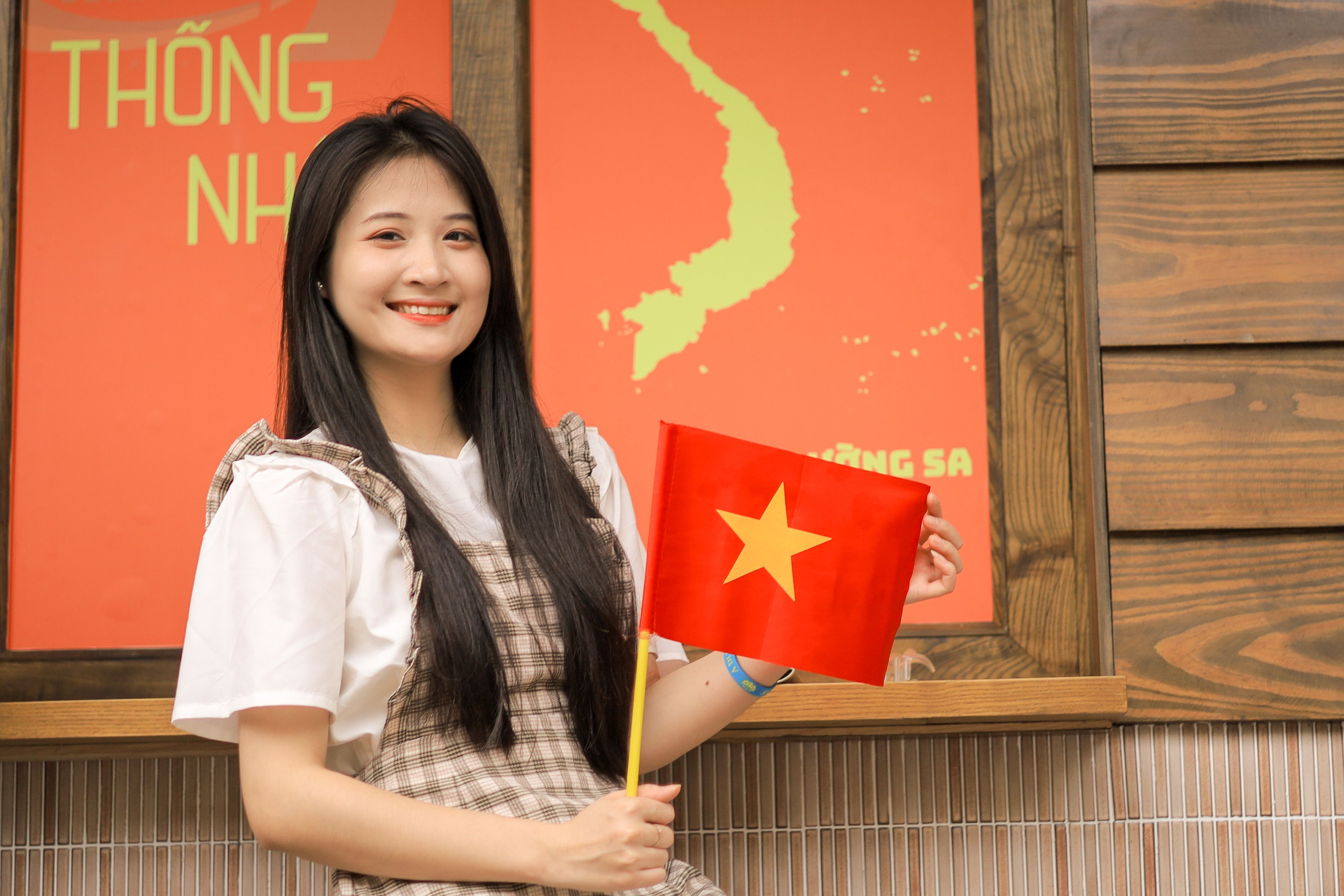 Young woman smiling indoors while holding a Vietnamese flag with a map in the background.