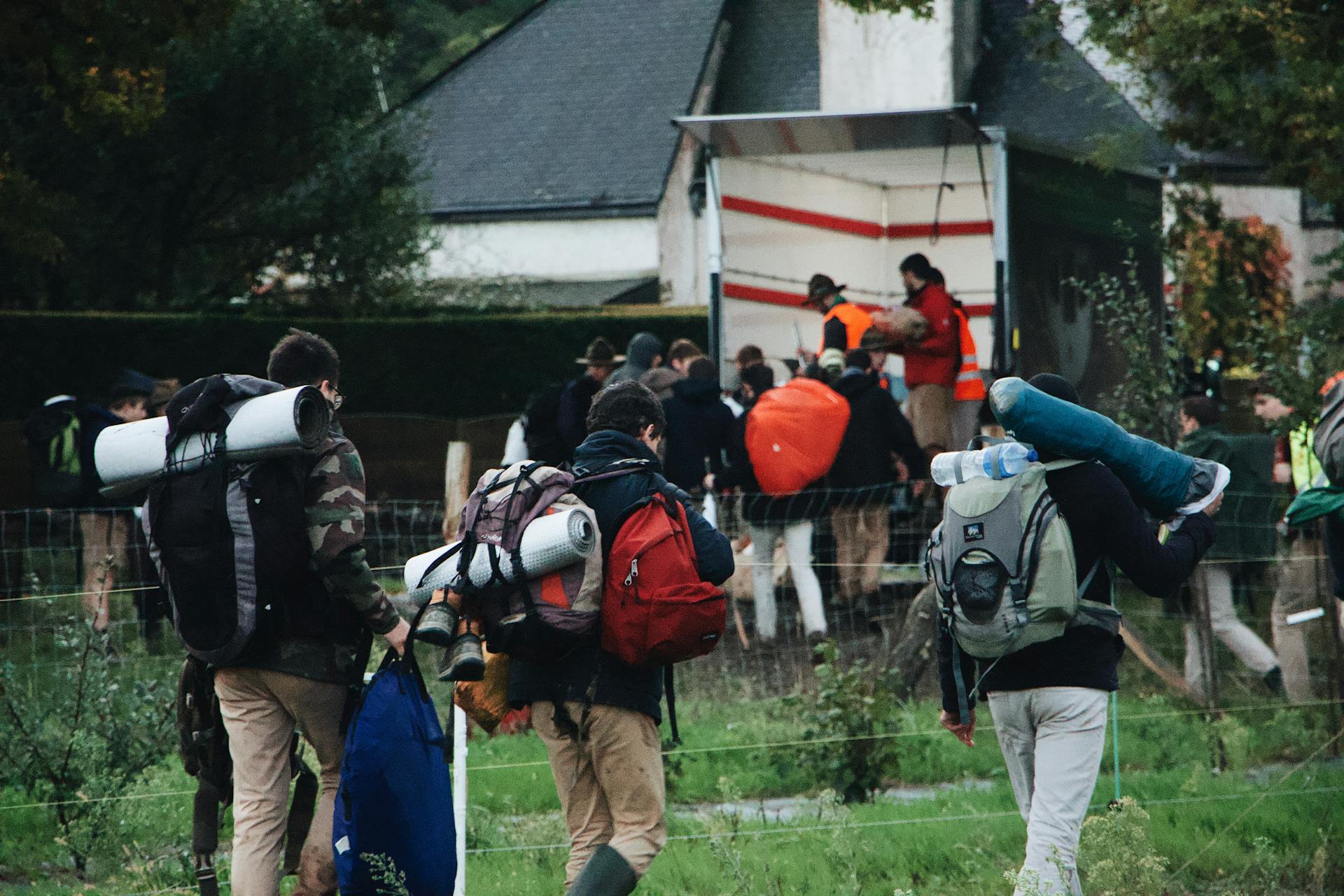 A group of people carrying backpacks and camping gear heading to a camp truck in a green outdoor setting.