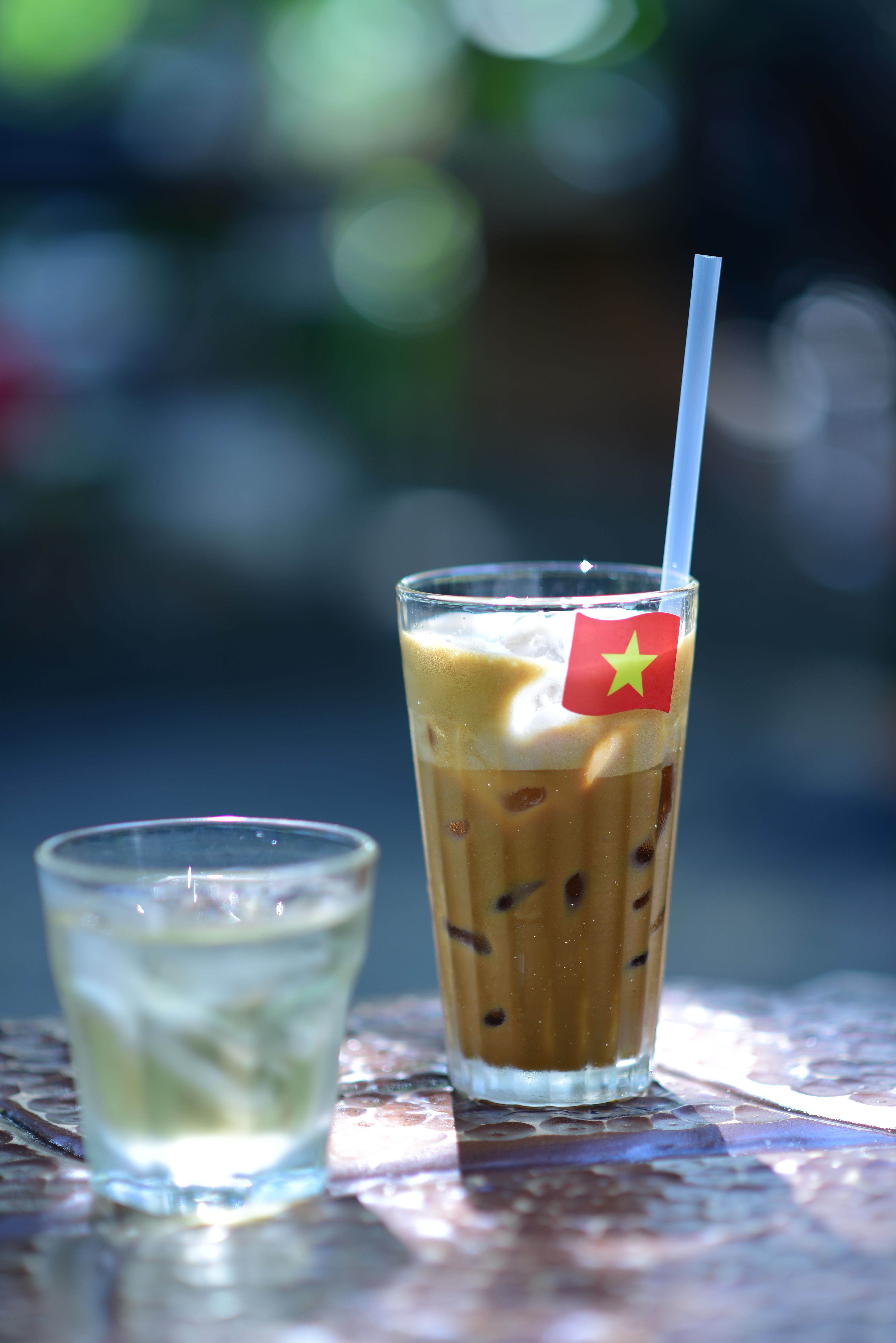 Glass of iced coffee with Vietnamese flag on a sunny outdoor table, next to a water glass.