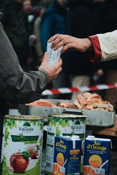 Hands exchanging a drink outdoors, surrounded by juice cartons and bread, evoking a communal sharing atmosphere.