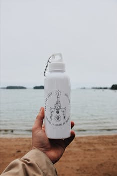 Hand holding a water bottle by the sea on a sandy beach, with a calm background.