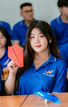 Young woman in classroom holding a red paper airplane, wearing a blue uniform, surrounded by classmates.