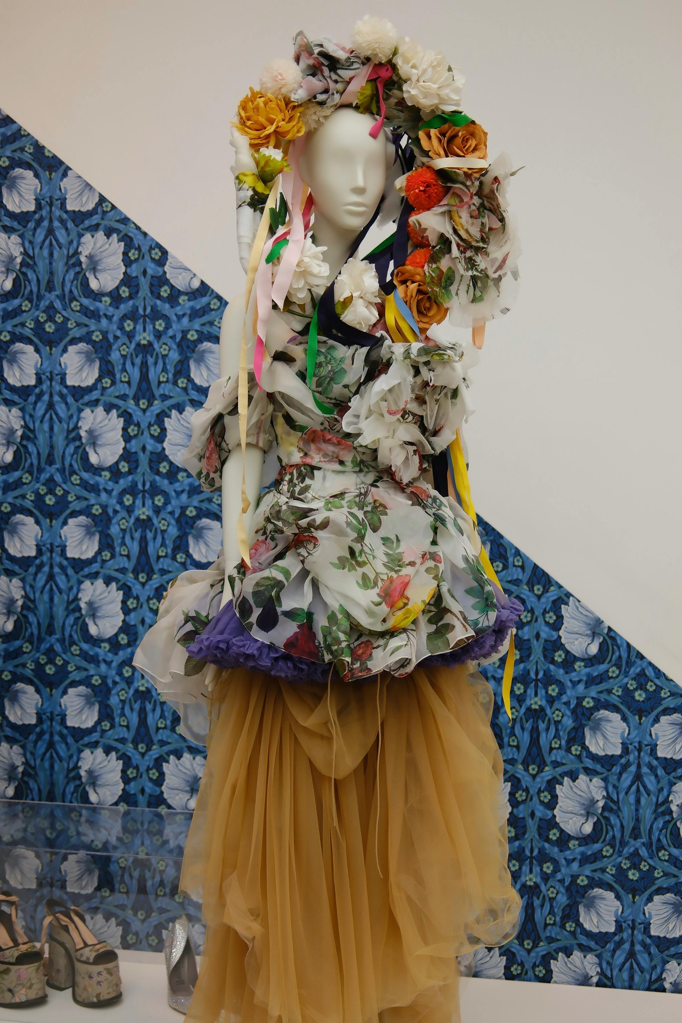 Free Mannequin dressed in vibrant floral attire with ornate headpiece and ribbons on a patterned backdrop. Stock Photo