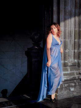 A woman in a flowing blue dress poses elegantly against a textured stone wall in daylight.