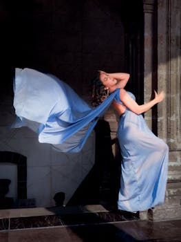 A woman in a flowing blue dress poses elegantly indoors, showcasing beauty and grace.
