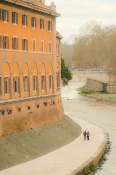 Couple strolls along a riverside path, framed by rustic architecture and soft light.