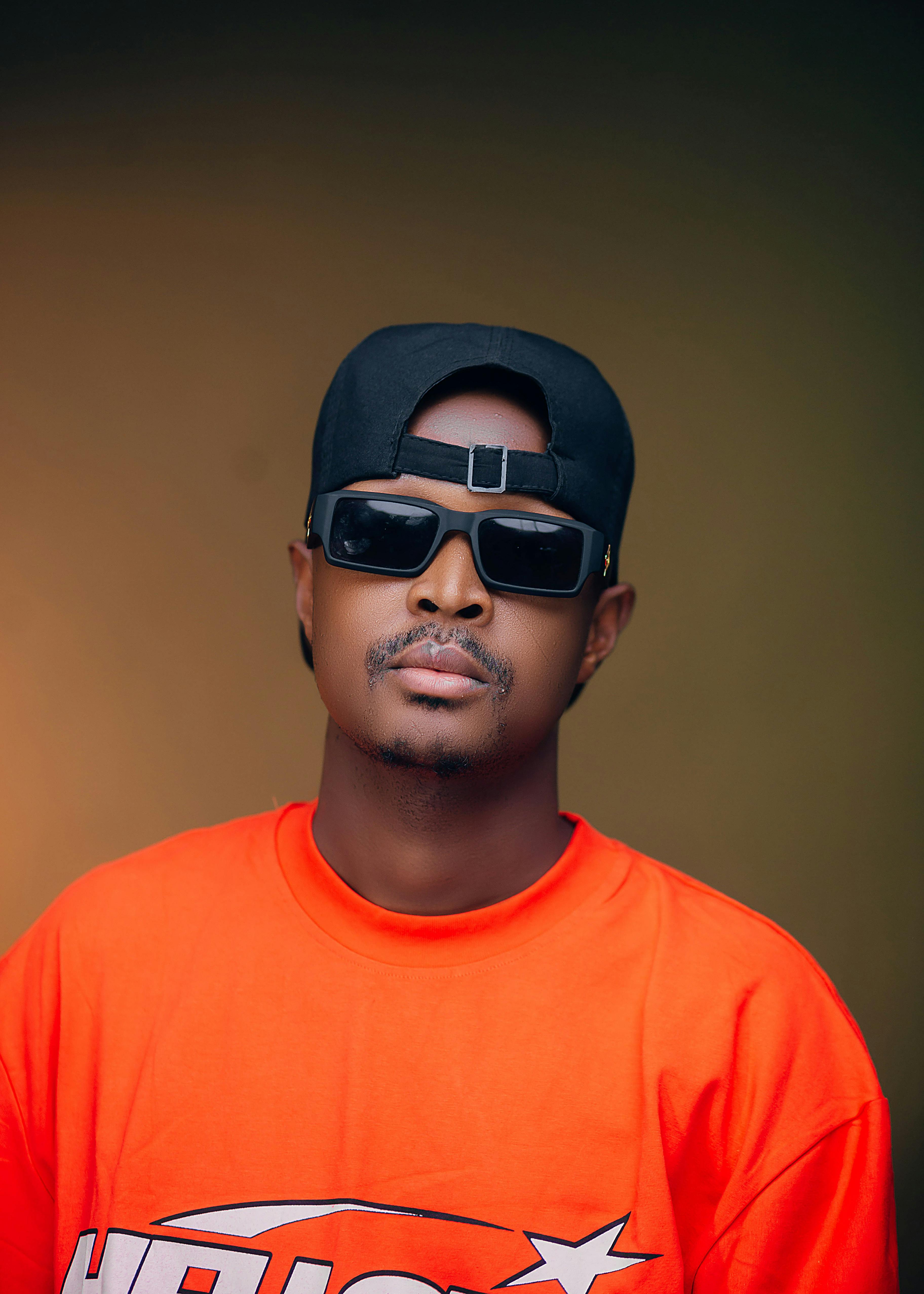 Cool portrait of a man wearing sunglasses, a cap, and an orange shirt against a smooth background.