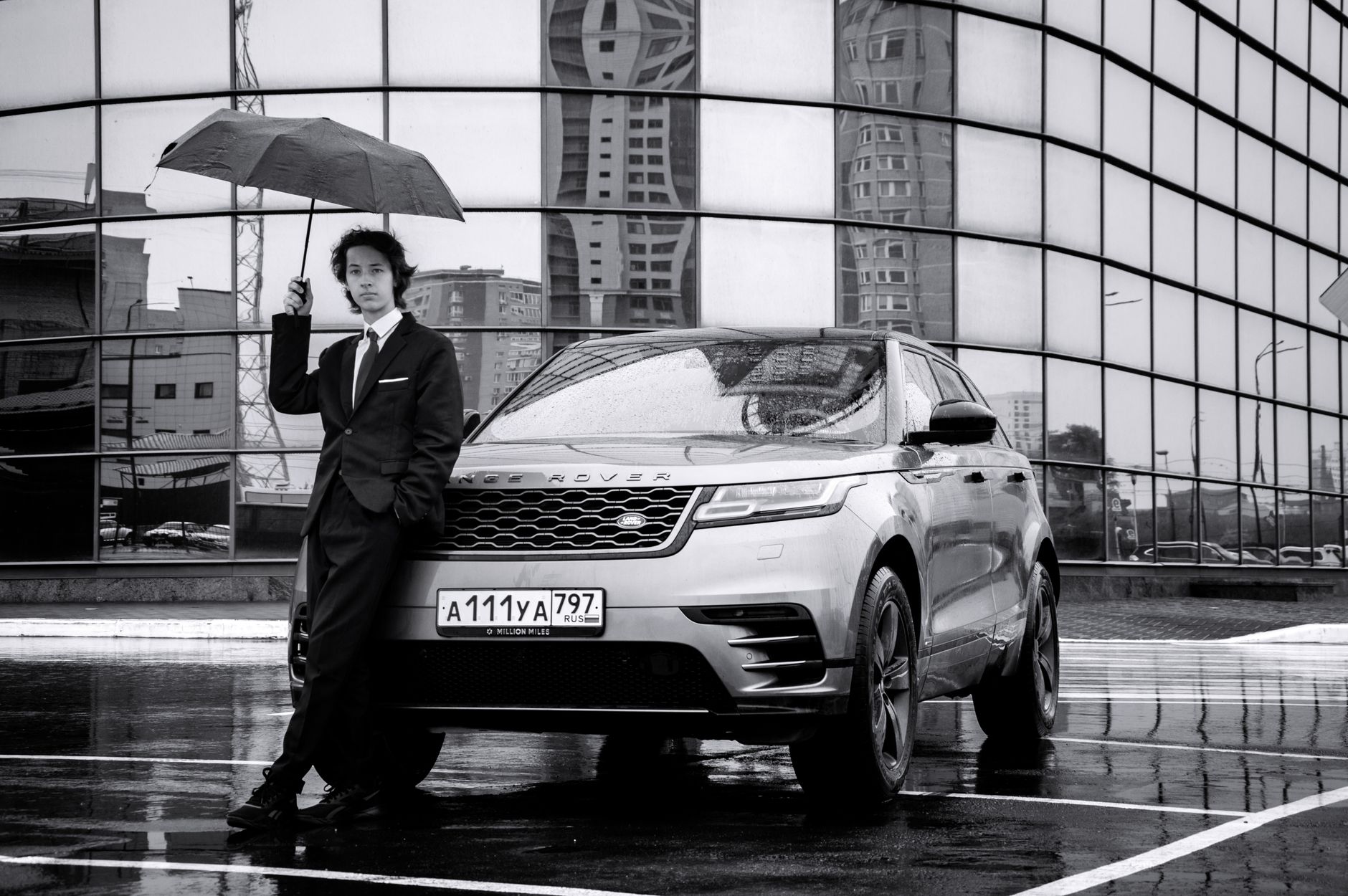Man in suit with an umbrella beside a Range Rover in the city, modern glass architecture.