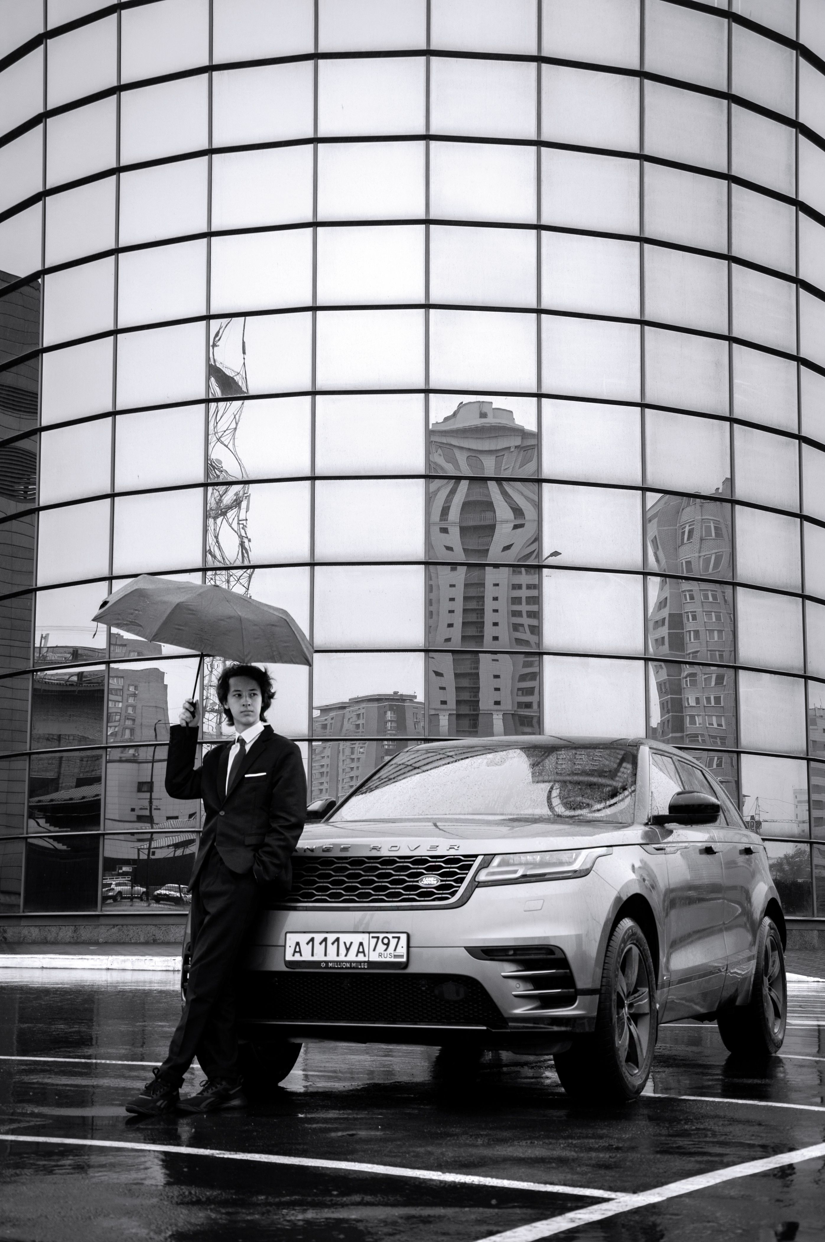 Monochrome image of a stylish man with umbrella next to a luxury car in a city setting.