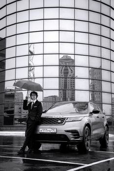 Monochrome image of a stylish man with umbrella next to a luxury car in a city setting.
