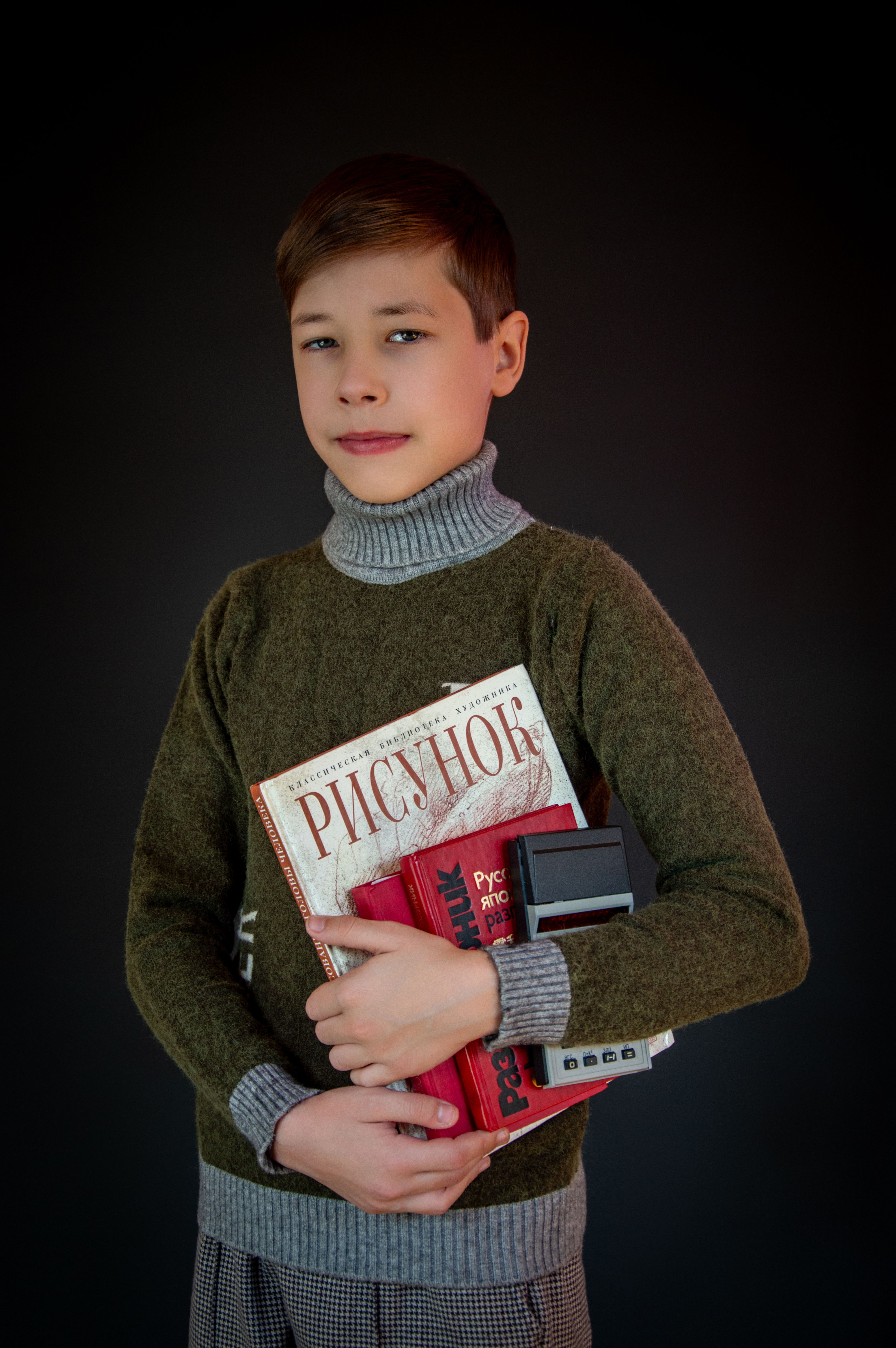 Portrait of young boy holding books, including one with Cyrillic text, against a dark backdrop.