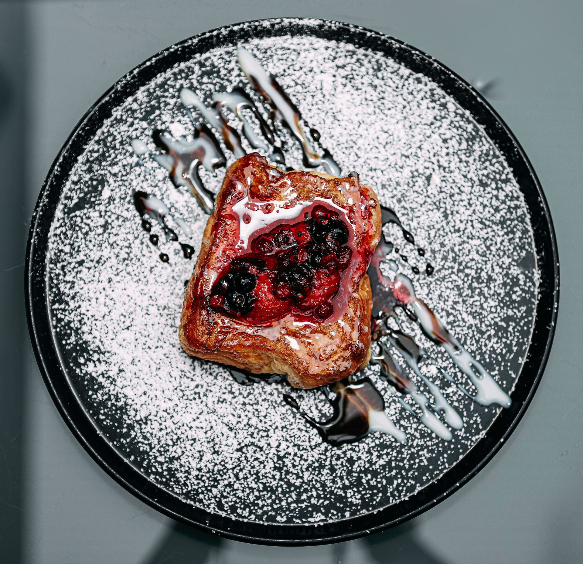 Aerial view of gourmet French toast topped with mixed berries and drizzled with syrup on a black plate.