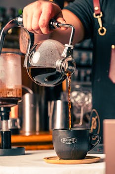 Barista skillfully pours hot brewed coffee from a glass pot into a cup in a cozy café setting.