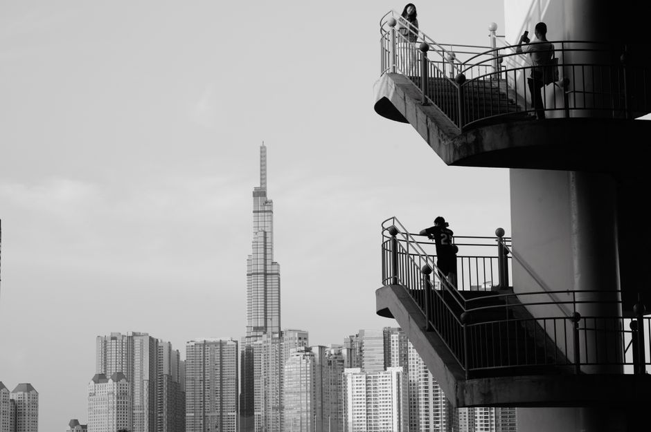 Black and white cityscape with people on a spiral staircase, modern skyscrapers in background.