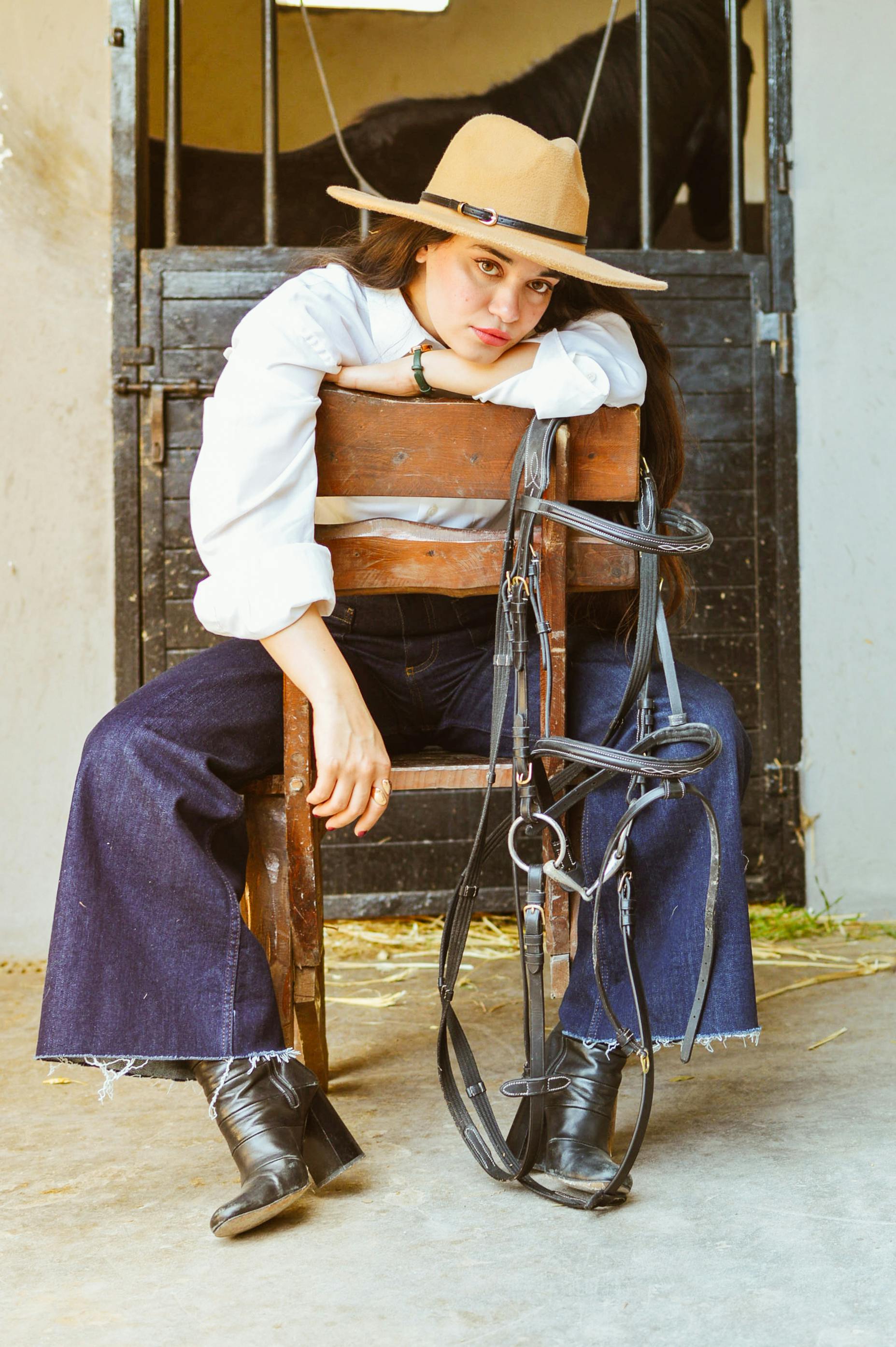 A fashionable young woman in a hat seated on a chair in a barn with horse tack.