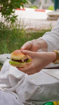 A close-up view of hands holding a fresh burger outdoors during daytime.