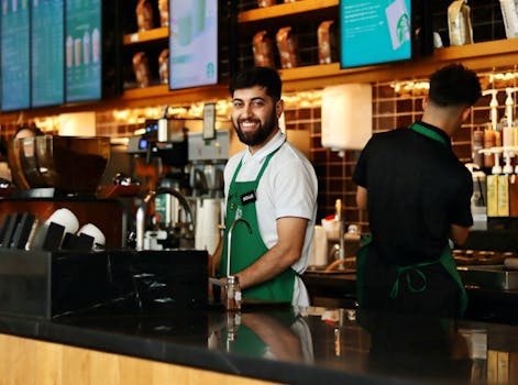 Friendly barista in a green apron smiling at the camera while working behind the coffee shop counter.