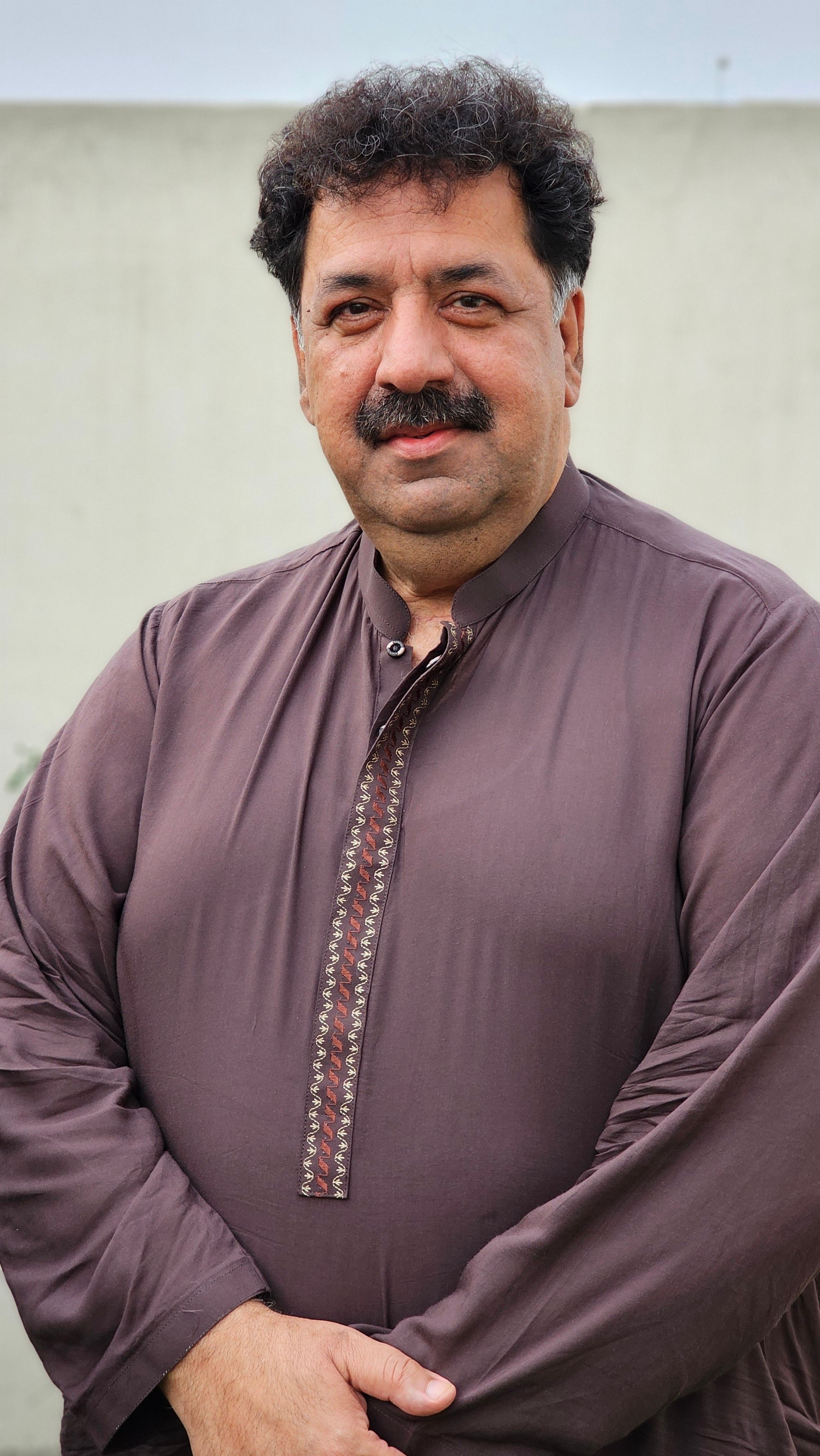 A confident man poses outdoors in traditional brown attire against a neutral background.
