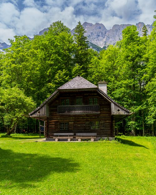 Free A serene log cabin surrounded by green forests and mountains under a cloudy sky. Stock Photo
