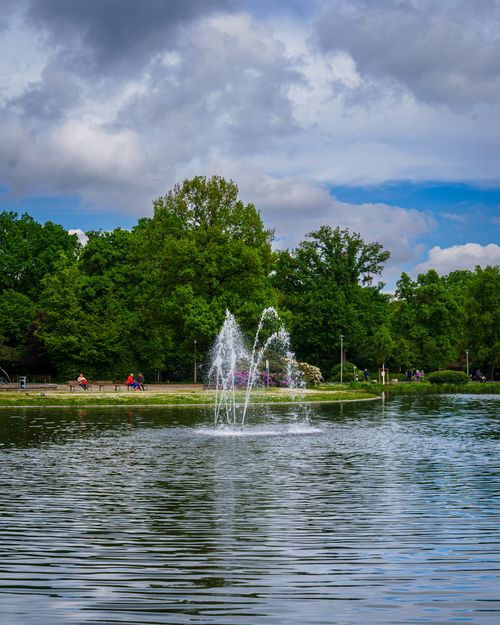 Free A tranquil park scene with a pond and water fountain surrounded by lush greenery under a cloudy sky. Stock Photo