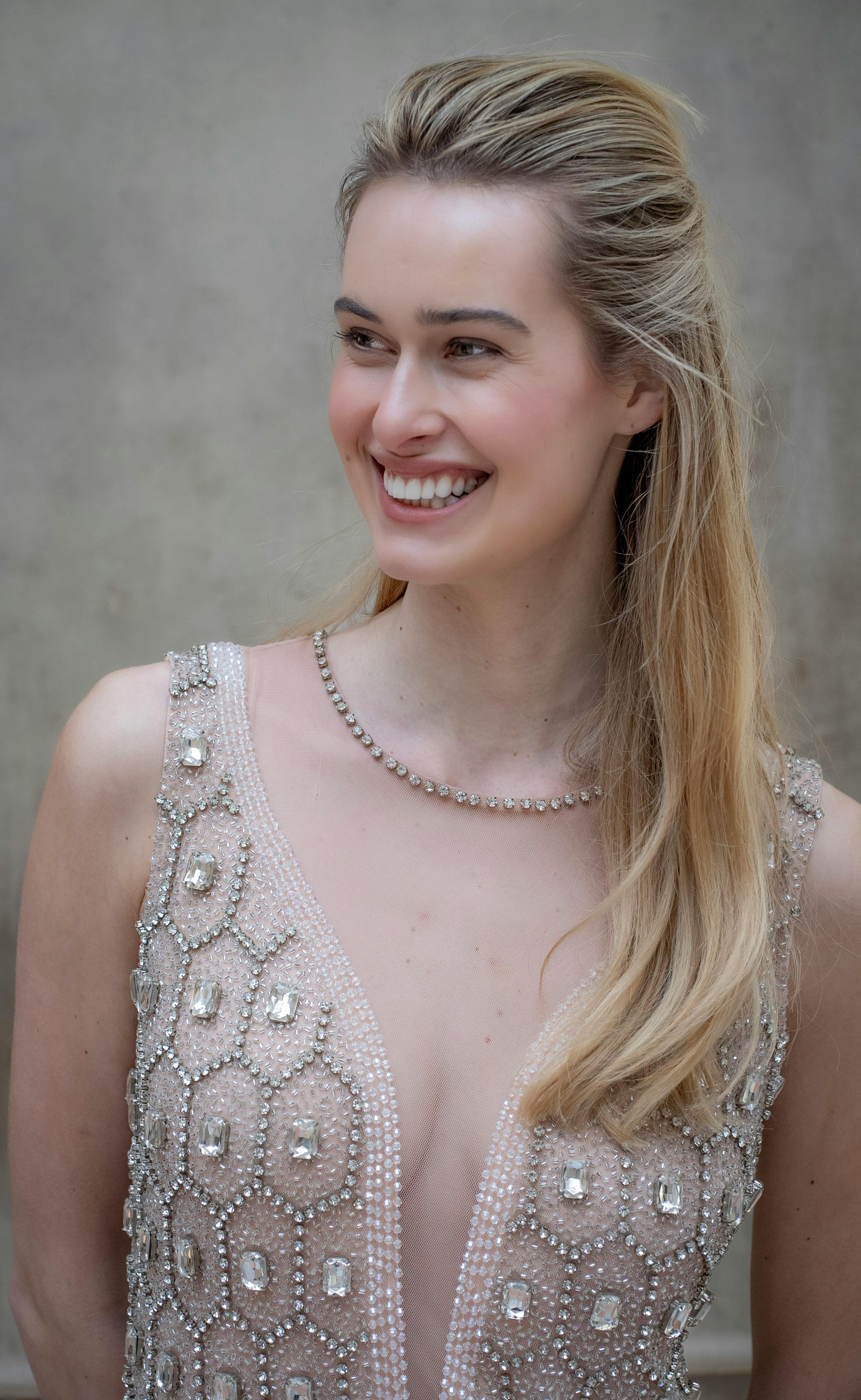 Smiling blonde woman in an intricate beaded dress against a neutral background, Milan.