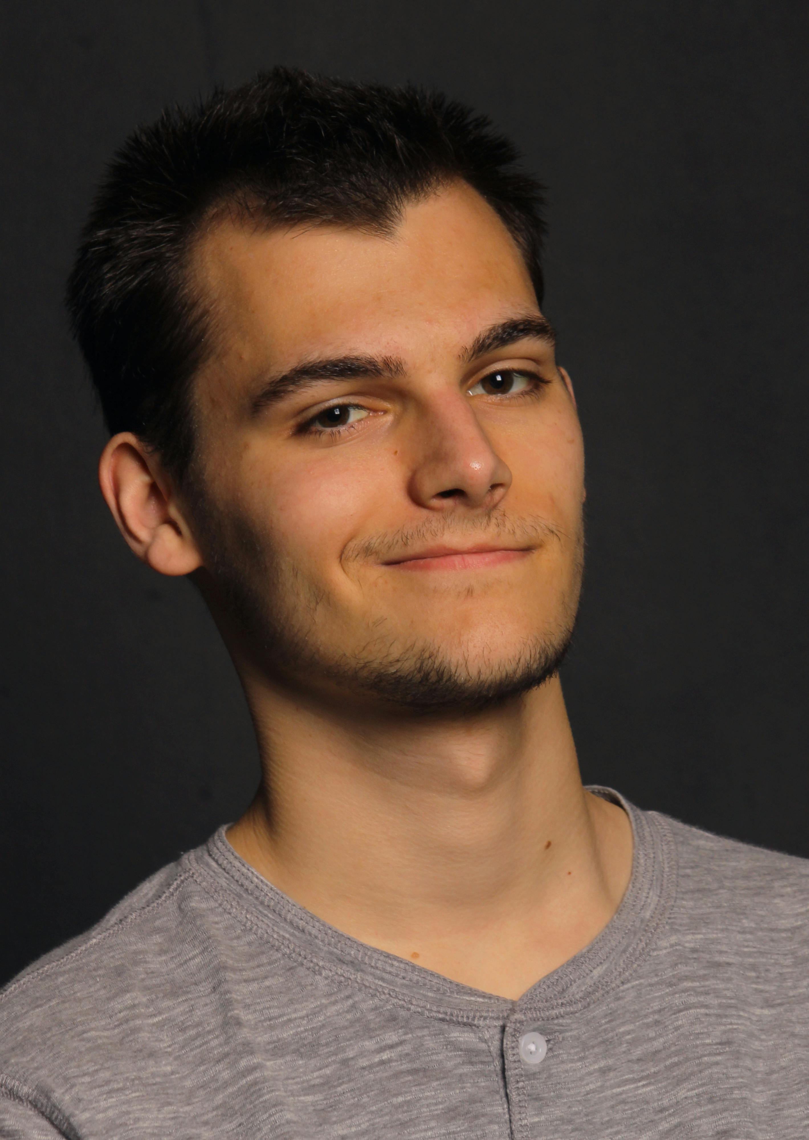 Close-up portrait of a young man smiling, set against a plain dark background.