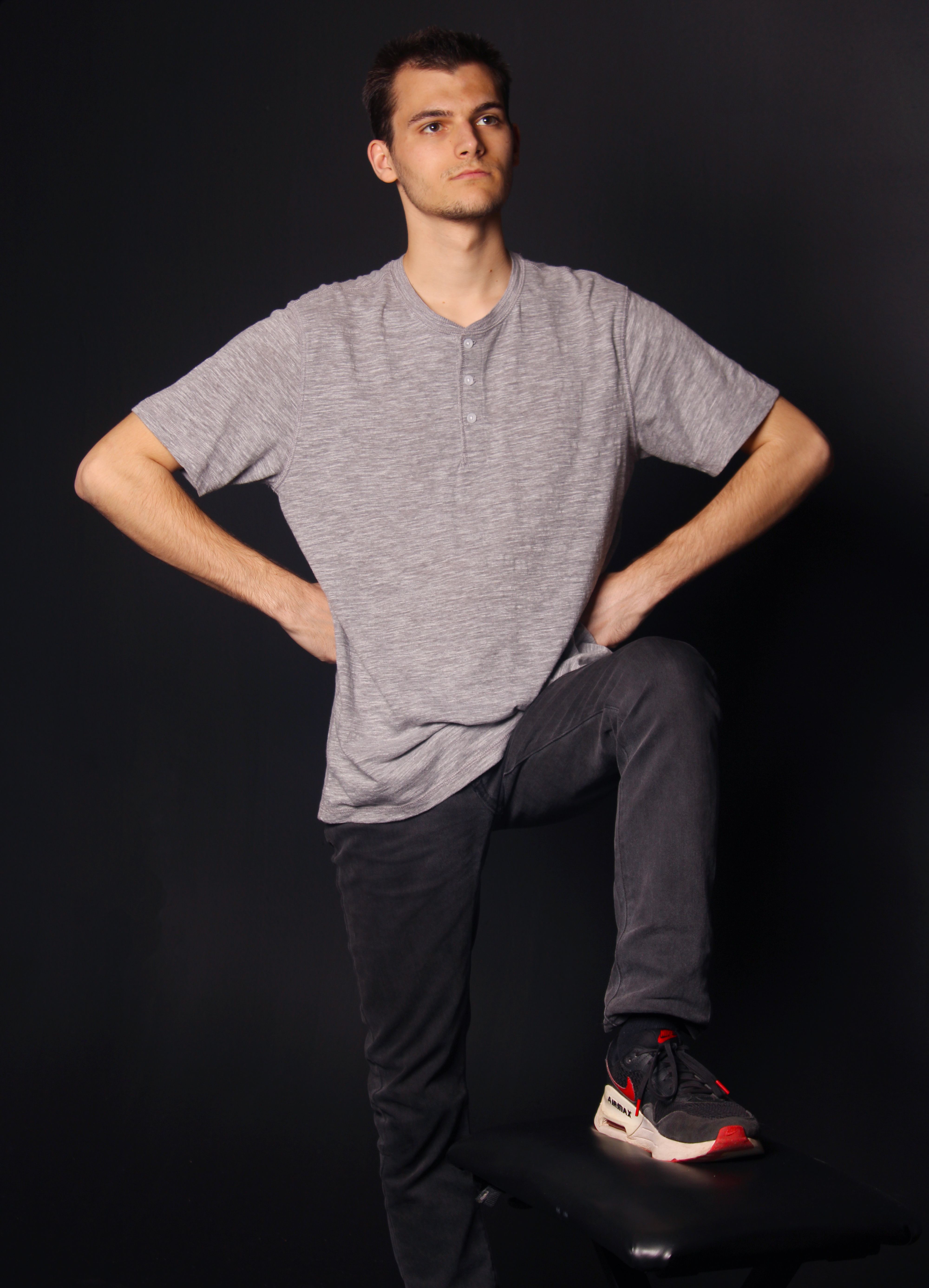 Young man in casual attire confidently posing with one foot on a stool against a black background.