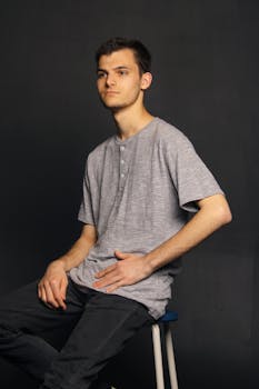 Portrait of a young man in a casual grey shirt sitting on a stool against a dark backdrop.