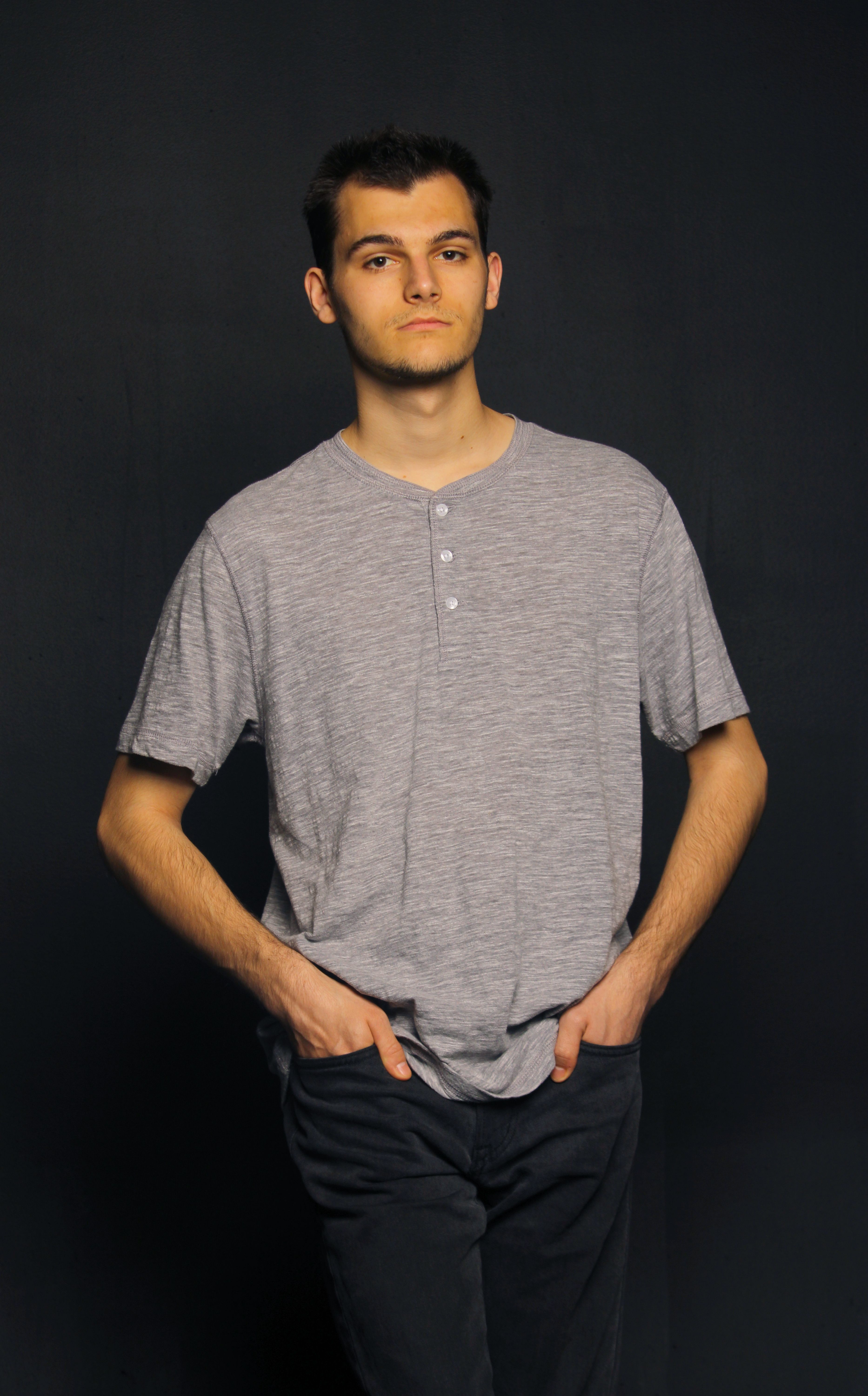 Portrait of a young man in a casual gray shirt with hands in pockets against a dark background.