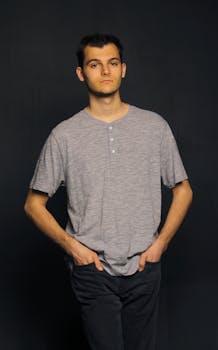 Portrait of a young man in a casual gray shirt with hands in pockets against a dark background.
