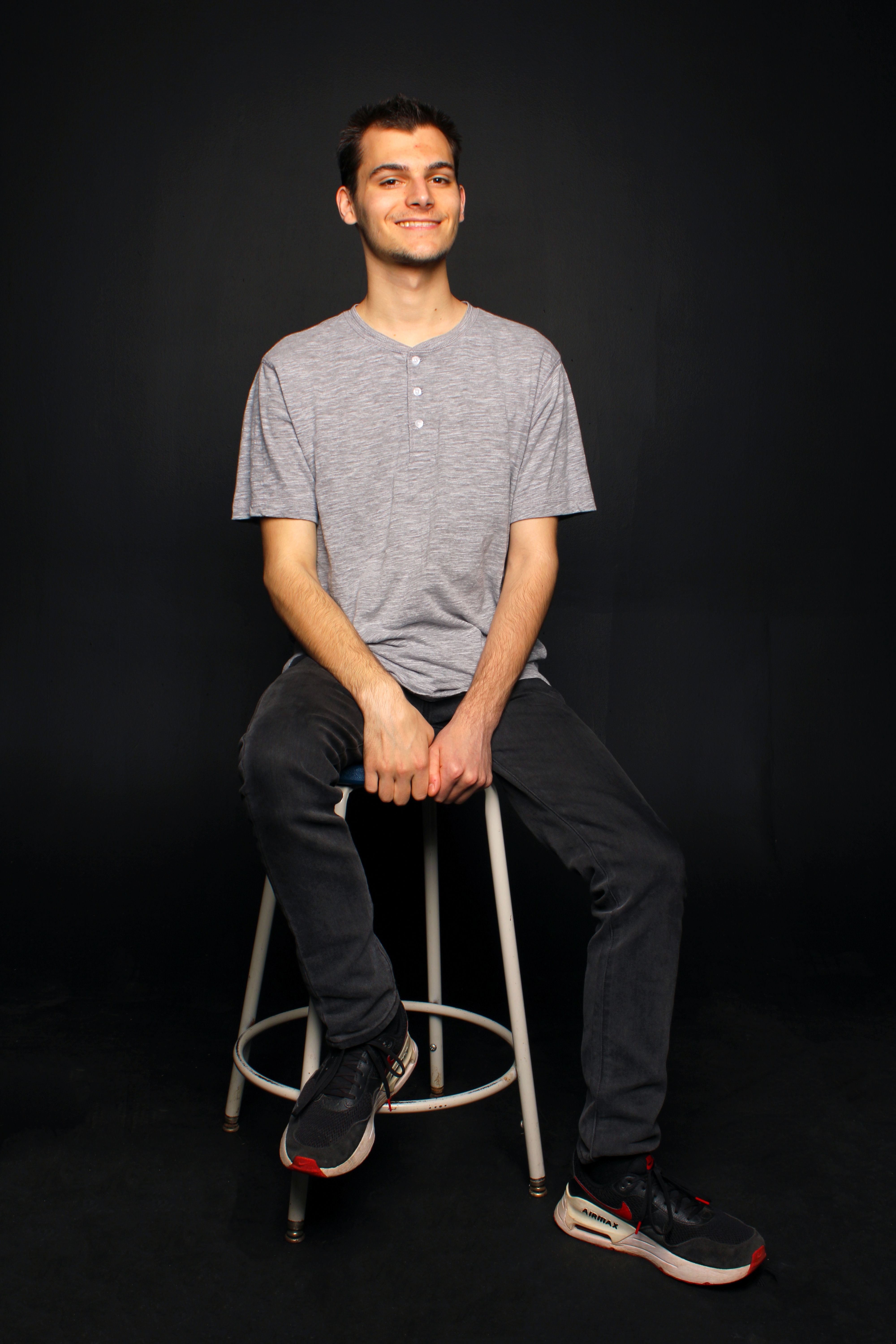 A relaxed man in a gray shirt and jeans sits smiling on a stool against a black backdrop.
